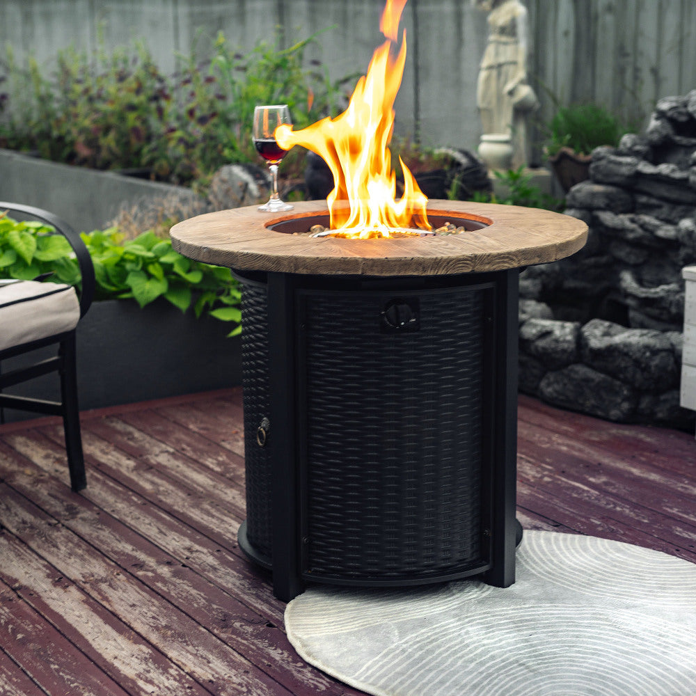 Round fire pit table with wooden top and black woven base, flames burning with wine glass nearby on deck.