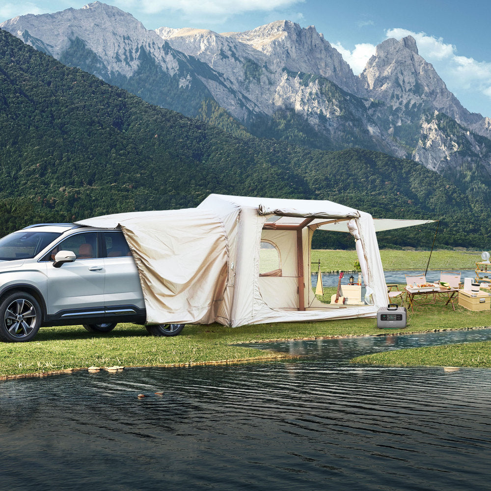 Beige canopy tent attached to an SUV, set up by a serene lake with mountain backdrop, creating an inviting outdoor space.