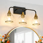 Black & gold industrial 3-light vanity wall sconce with clear glass shades above a round gold-framed mirror with floral decor in foreground.