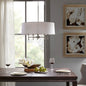 5-light drum shade chandelier with white cotton-linen shade and silver plated brass frame, hanging over dark wood dining table with plates, wine glasses, and floral centerpiece.