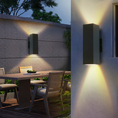 Two modern black outdoor wall sconces with up & down lighting mounted on patio wall, illuminating wooden dining table and chairs in evening setting.