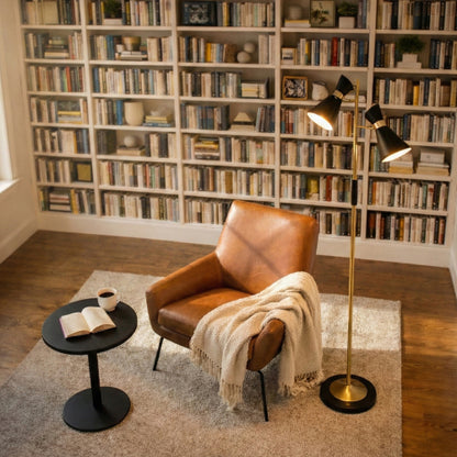 Double spot floor lamp with brushed gold finish and metal base, 4-way switch, beside a leather armchair with a coffee cup and open book in a library reading nook.