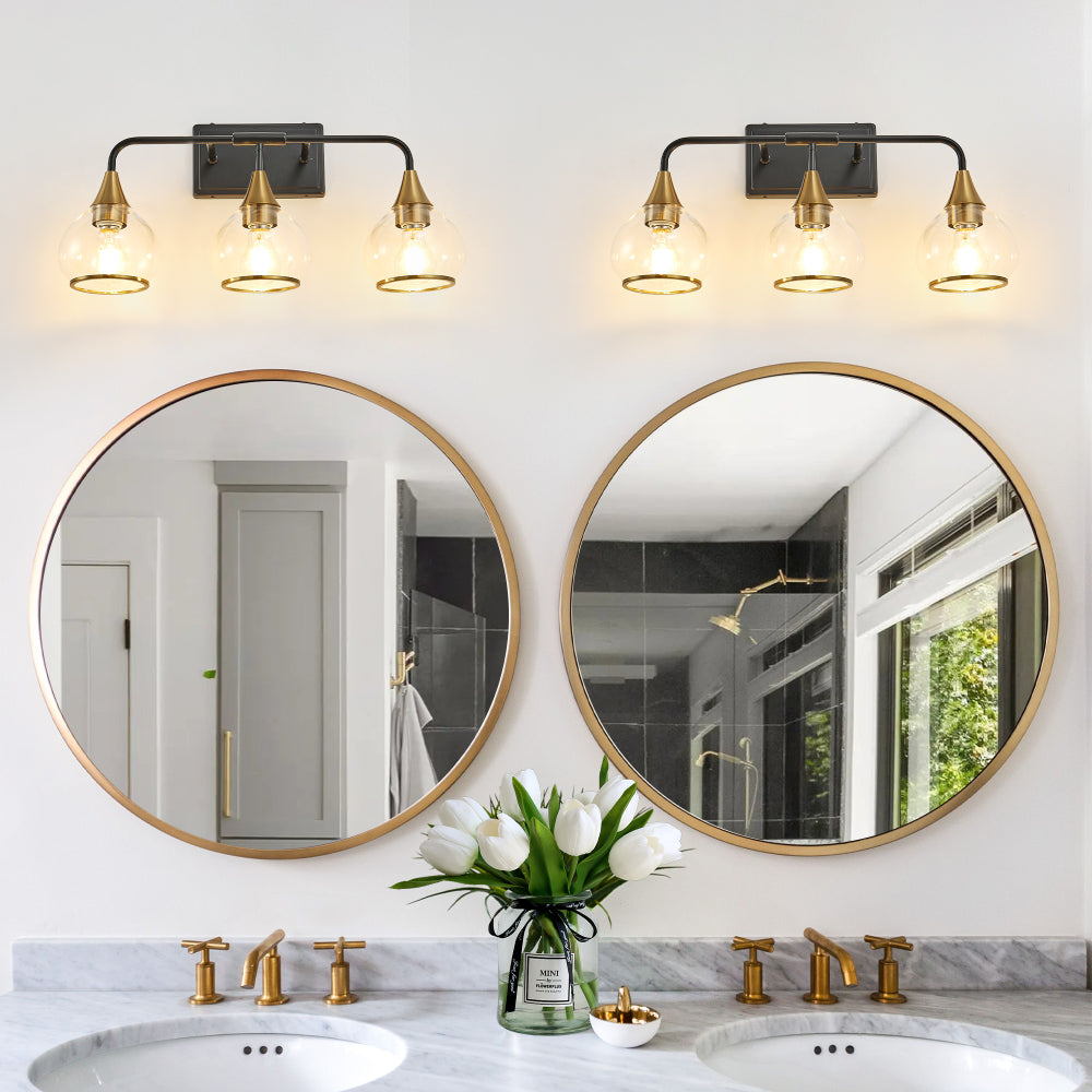 Black & gold industrial vanity wall sconces with clear glass shades, 3/4-light fixtures above double round mirrors in a bathroom with marble countertop and gold faucets.