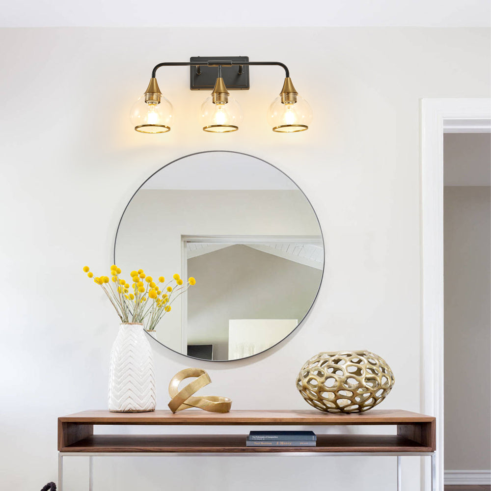 Black & gold industrial 3-light vanity wall sconce with clear glass shades above a round mirror in a living space with console table and yellow floral decor.