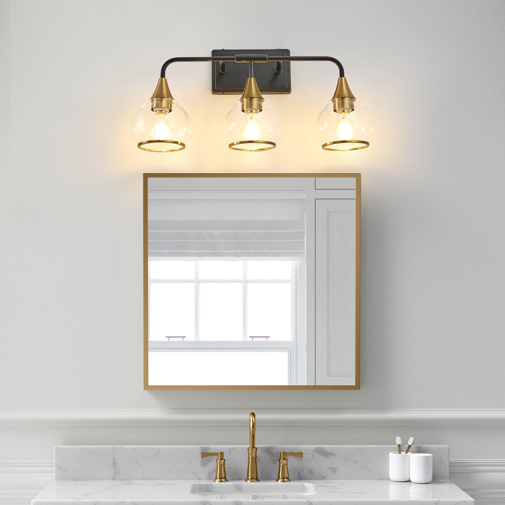 Black & gold industrial 3-light vanity wall sconce with clear glass shades above a rectangular gold-framed mirror in a bathroom with marble countertop and gold faucet.