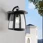 Black solar wall lantern with clear glass panels mounted on light gray wall, featuring dual solar panels and 4 lighting modes, with white house and blue sky in background