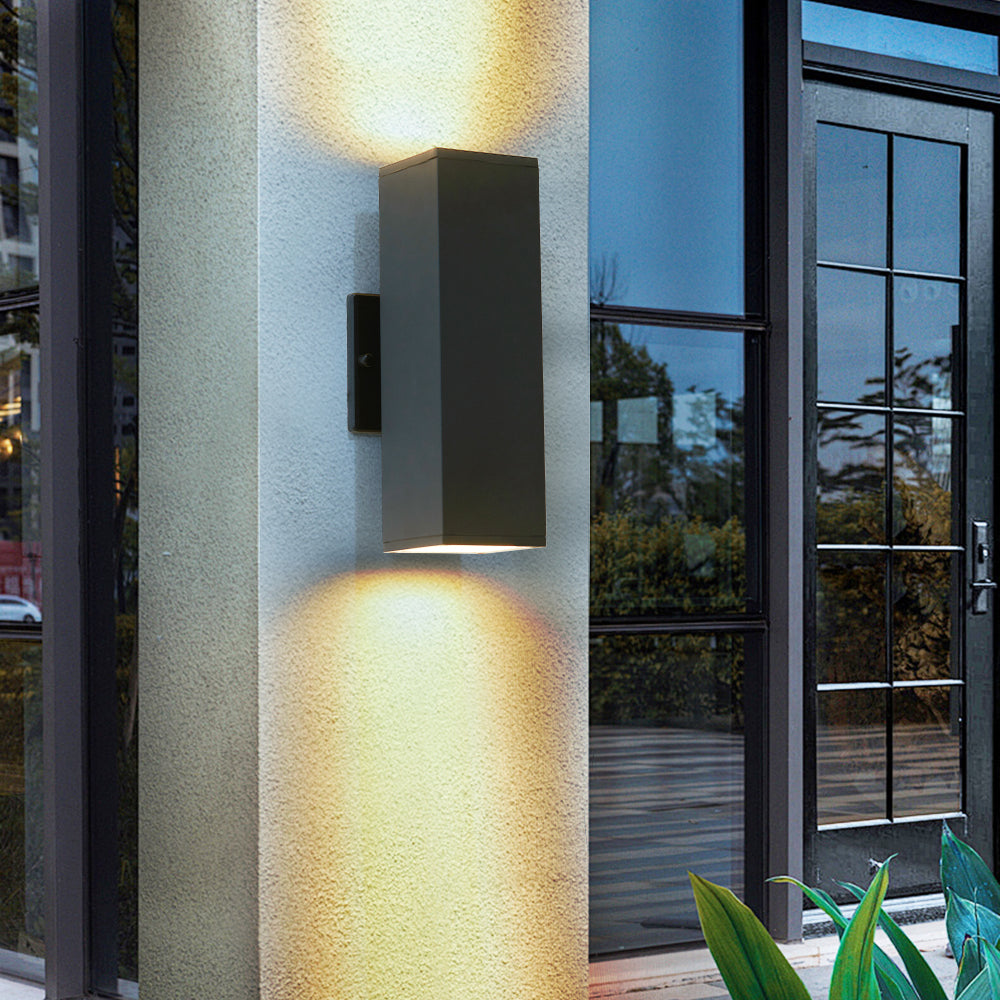 Modern black outdoor wall sconce with up & down lighting mounted on exterior column, casting warm glow on wall near glass door.