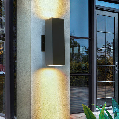Modern black outdoor wall sconce with up & down lighting mounted on exterior column, casting warm glow on wall near glass door.