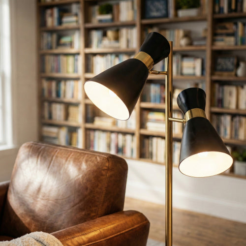 Modern double spot floor lamp with brushed gold metal base and black adjustable shades, providing reading and accent lighting in a cozy living space with bookshelves.