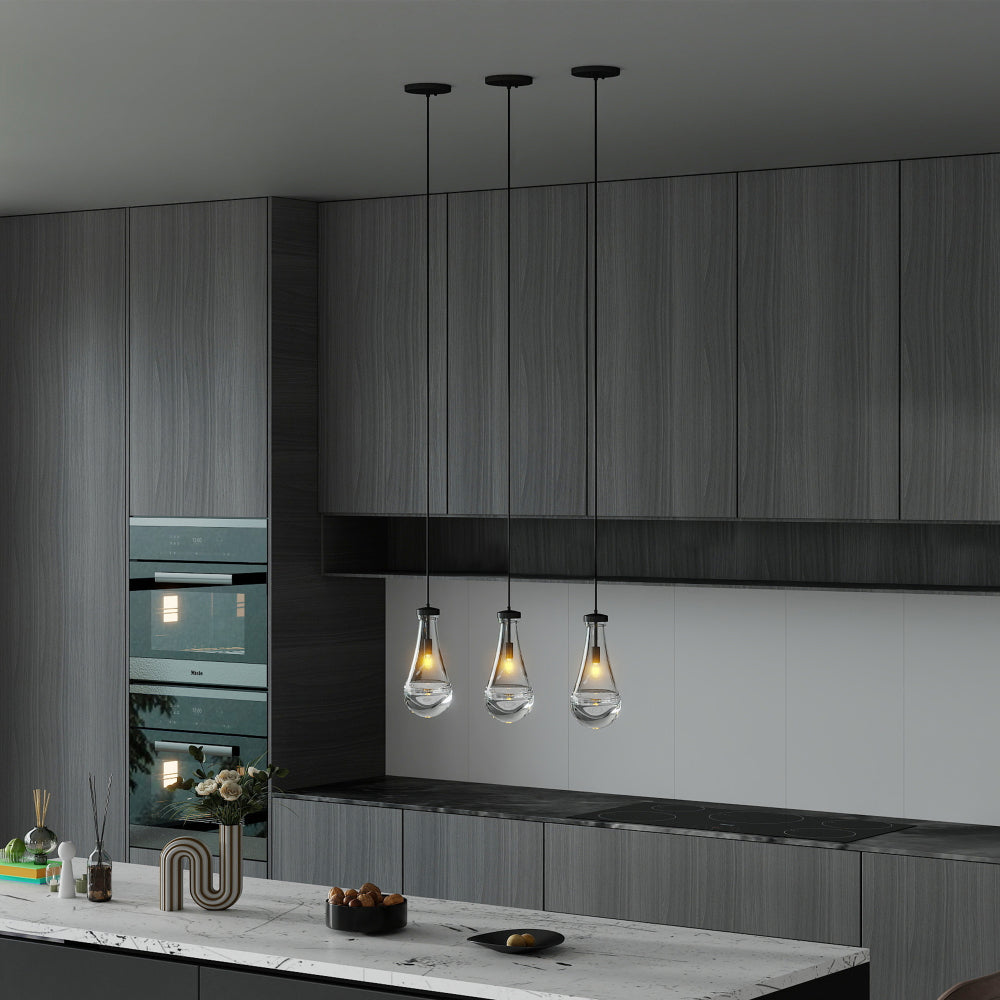 Three Raindrop Pendant Lights with hand-blown clear glass shades and brass/black rods hanging over a modern kitchen island, emitting warm ambient light.