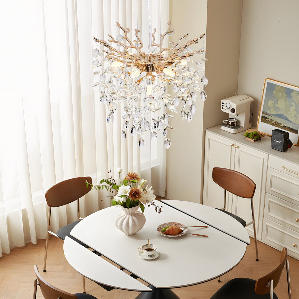 Gold tree branch crystal chandelier with crystal pendants over a white dining table with floral centerpiece and wooden chairs.