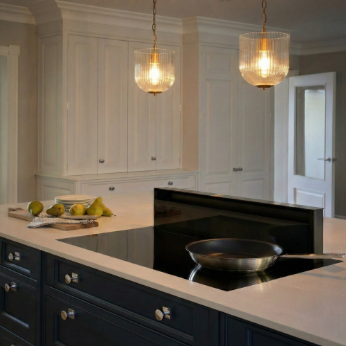 Two coarse gold metal pendant lamps with clear glass shades hanging over a kitchen island with white cabinets and a black cooktop.