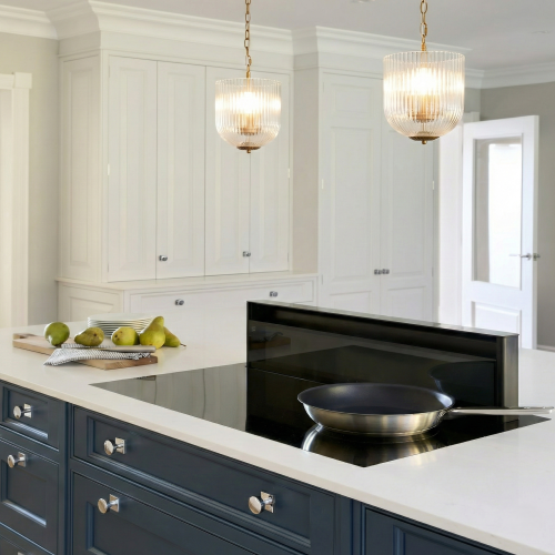 Two coarse gold metal pendant lamps with clear glass shades hanging over a kitchen island with white cabinets and a black cooktop.
