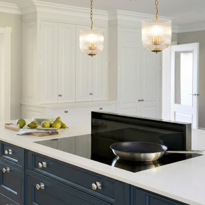 Two coarse gold metal pendant lamps with clear glass shades hanging over a kitchen island with white cabinets and a black cooktop.