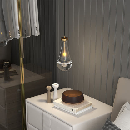Raindrop Pendant Light with hand-blown clear glass shade and gold rod, hanging above a nightstand with books and decor in a bedroom.