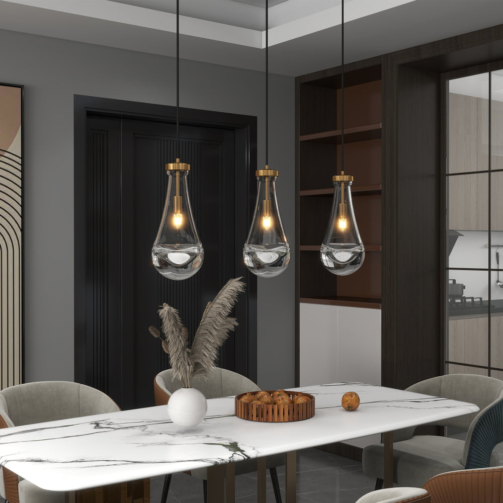 Three Raindrop Pendant Lights with hand-blown clear glass shades and brass accents, hanging over a marble dining table with pampas grass and bread basket.