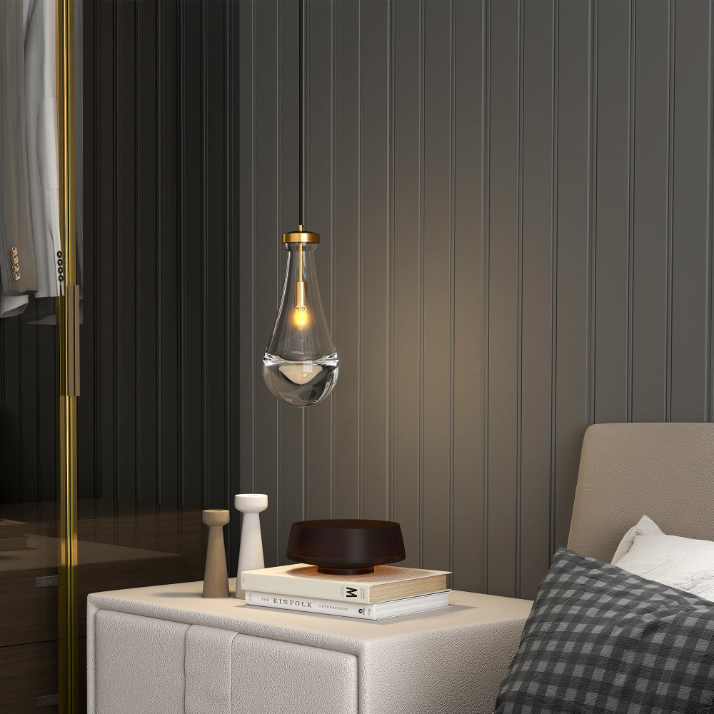 Raindrop Pendant Light: Hand-blown clear glass shade with brass rod, illuminated above a nightstand with books and decor in a bedroom.