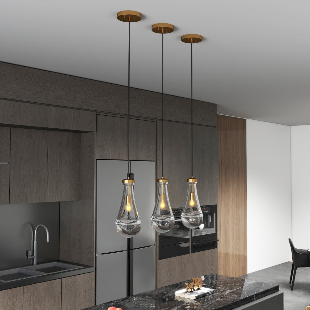Three Raindrop Pendant Lights with hand-blown clear glass shades and brass accents, hanging over a black marble kitchen island with dark wood cabinets and stainless steel appliances.