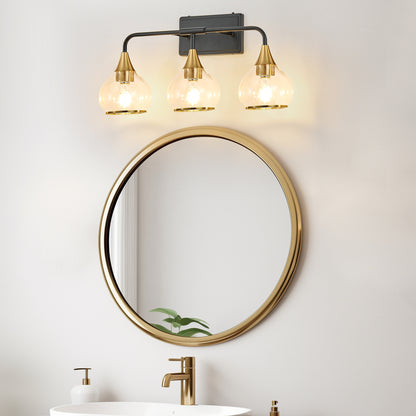 Black & gold industrial 3-light vanity wall sconce with clear glass shades above a round gold-framed mirror in a bathroom with marble sink and gold faucet.